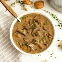 Overhead shot of bowl of best ever mushroom soup with a wooden handled spoon in the bowl on the left side of the bowl. Fresh mushrooms and thyme leaves surround the bowl and there is a grey striped napkin to the left of the bowl