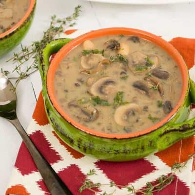 White Bean Mushroom Soup in a green and orange bowl with creamy mushrooms on the top, a red and orange napkin and a golden spoon next to the bowl