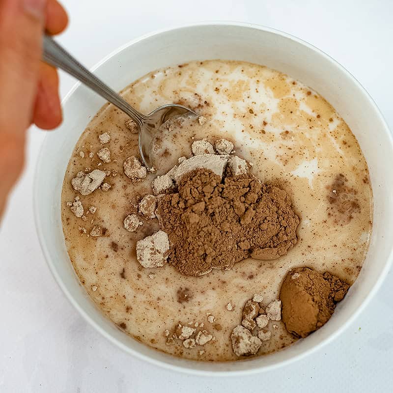 Protein powder, cocoa powder and milk in a bowl with a hand holding a spoon in the bowl.