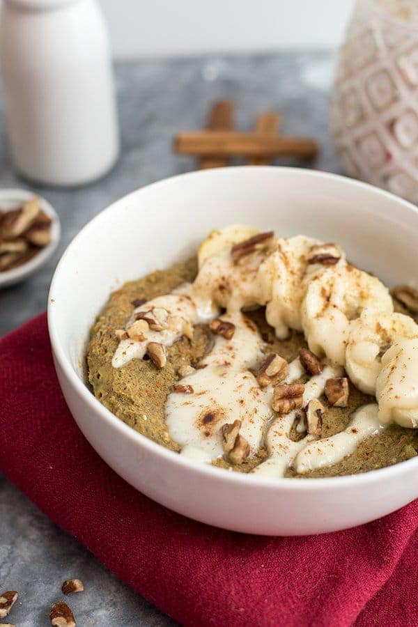 White bowl of gingerbread mug cake over a red napkin topped with sliced bananas, chopped pecans and coconut butter drizzle