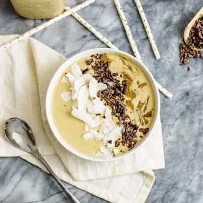 Peanut Butter Protein Smoothie in white bowl topped wtih coconut flakes, cacao nibs and peanut butter white jar in background