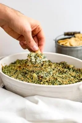 Hand dipping chip into spinach artichoke dip in a white casserole dish