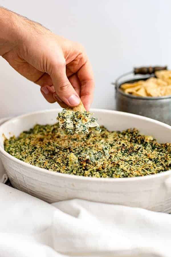 Hand dipping chip into spinach artichoke dip in a white casserole dish