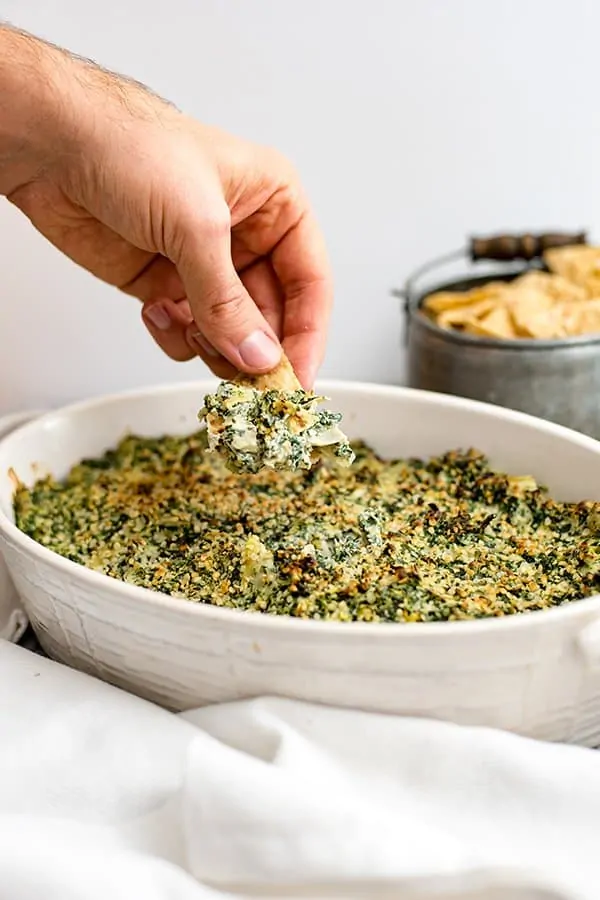 Hand dipping chip into spinach artichoke dip in a white casserole dish