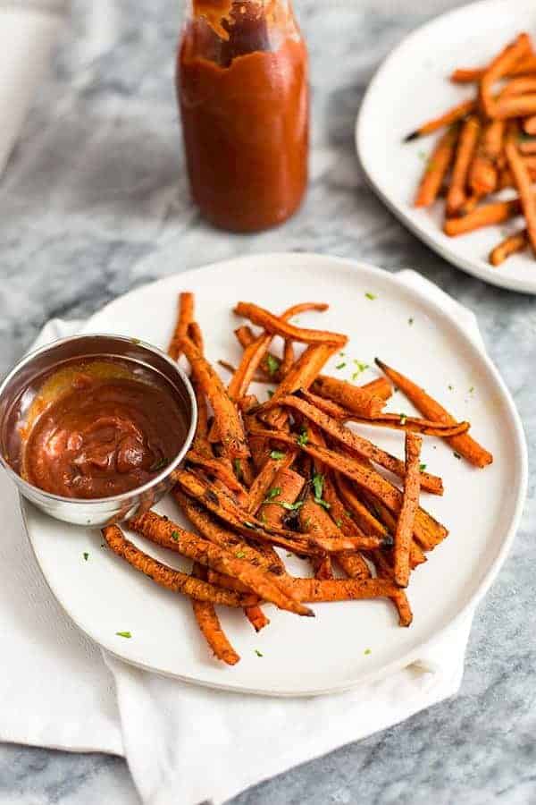 Carrot fries over a white plate with a side of ketchup in a silver bowl over a white napkin