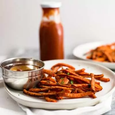 Carrot fries over a white plate with a side of ketchup in a silver bowl over a white napkin. Ketchup in the background