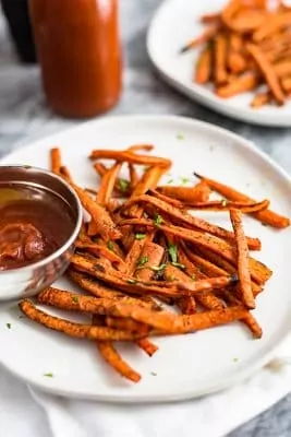 Carrot fries over a white plate with a side of ketchup in a silver bowl over a white napkin. Ketchup in the background