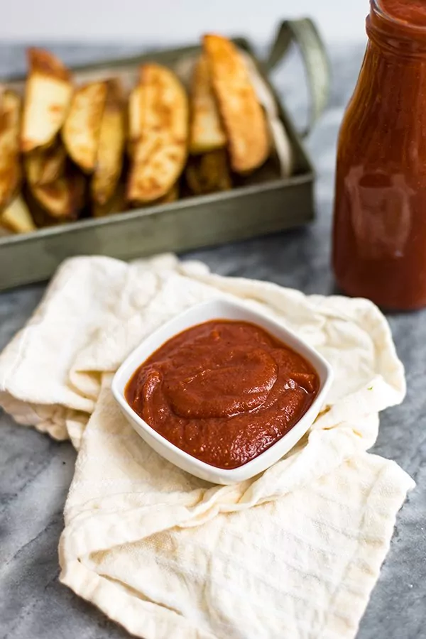 White ramekin filled with homemade ketchup on a napkin, a bottle of homemade ketchup and french fries in the background