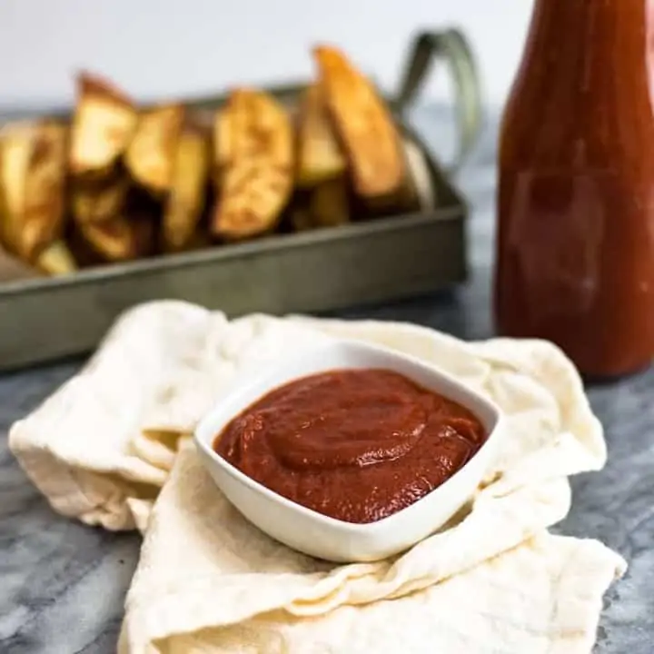 White ramekin filled with homemade ketchup in the center, a bottle of homemade ketchup and french fries in the background