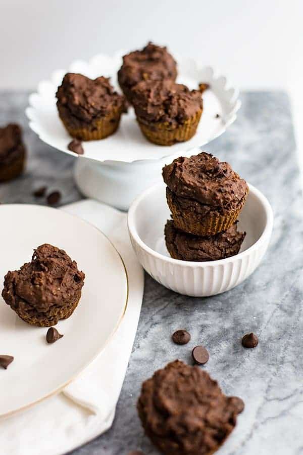 Vegan Chocolate Protein Muffins stacked in a white bowl surrounded by chocolate chips and other chocolate protein muffins
