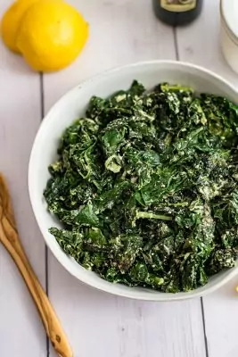 Overhead shot bowl of creamy kale with a lemon in the background and a wooden spoon to the left