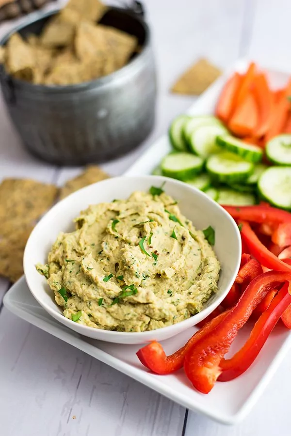 Bowl of roasted cauliflower hummus with sliced red bell pepper, cucumber and carrots and basket of chips 