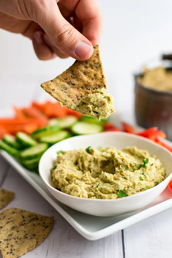 chip being dipped into roasted cauliflower hummus with vegetables in the background