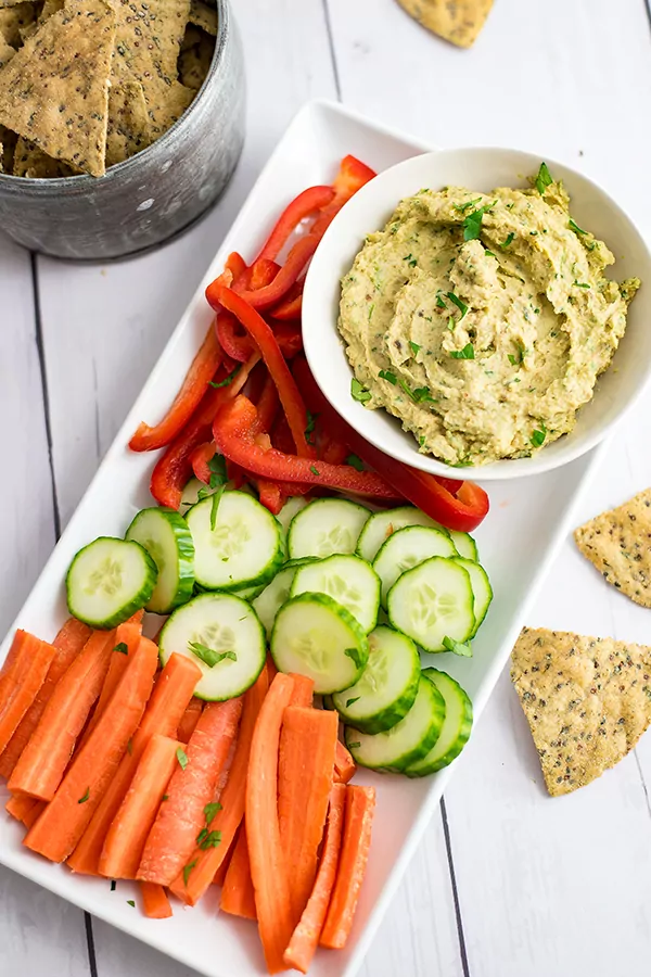 White platter with chopped carrots, cucumber and red bell pepper along a bowl of roasted cauliflower hummus