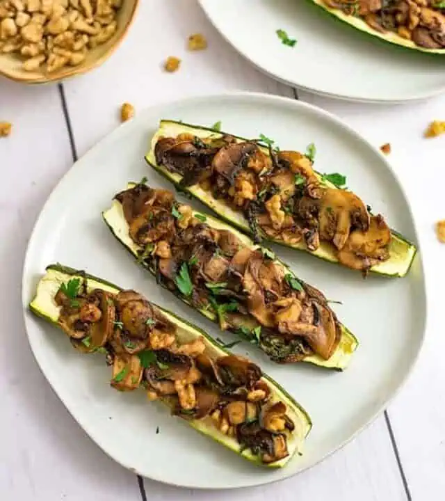 Three stuffed zucchini on an off white plate with a bowl of chopped walnuts in the background