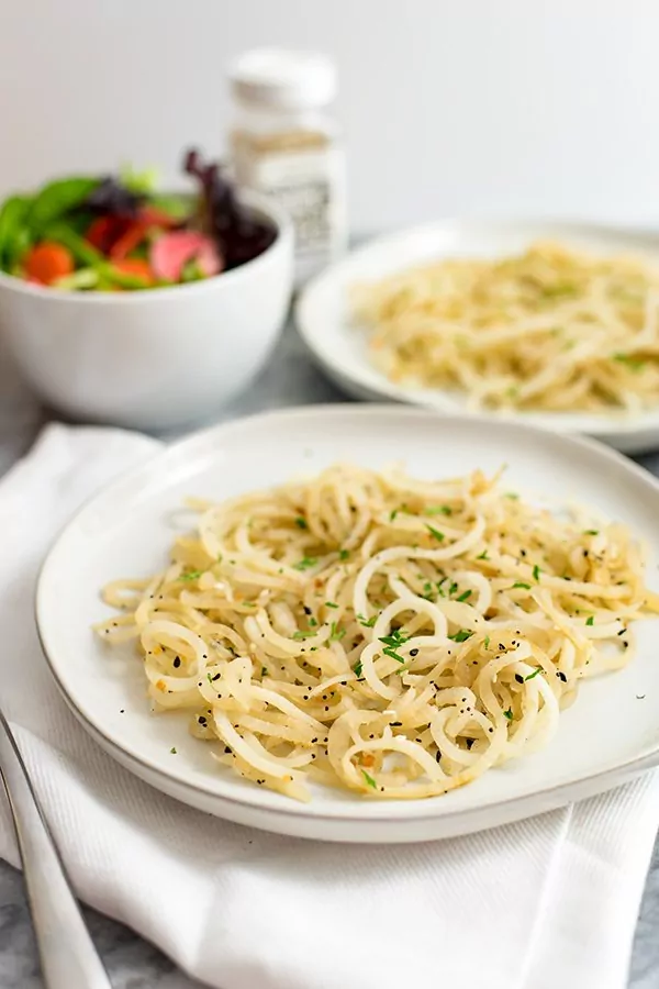 white bowl with low carb pasta made of daikon radish on a white napkin with parsley on top with a salad in a white bowl and a second plate of low carb pasta off to the right in the background