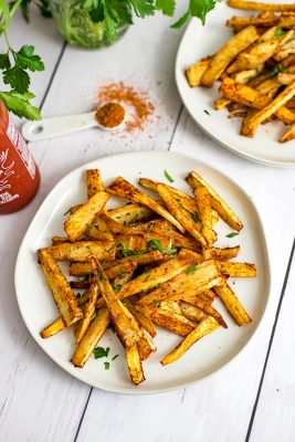 Cajun parsnip fries on a white plate with a teaspoon of cajun seasoning and an additional plate of parsnip fries in the background