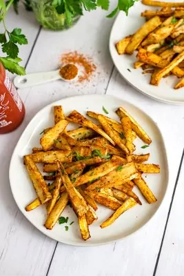 Cajun parsnip fries on a white plate with a teaspoon of cajun seasoning and an additional plate of parsnip fries in the background