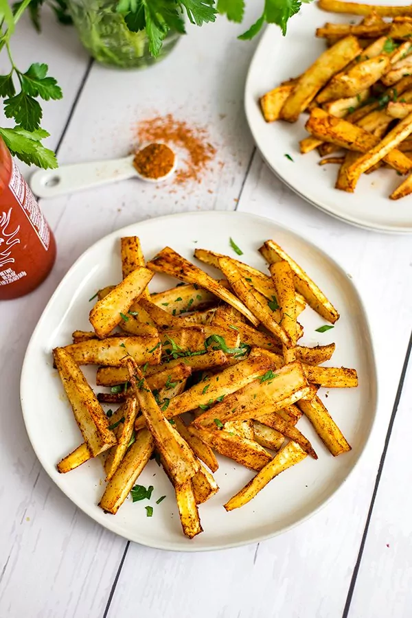 Cajun parsnip fries on a white plate with a teaspoon of cajun seasoning and an additional plate of parsnip fries in the background