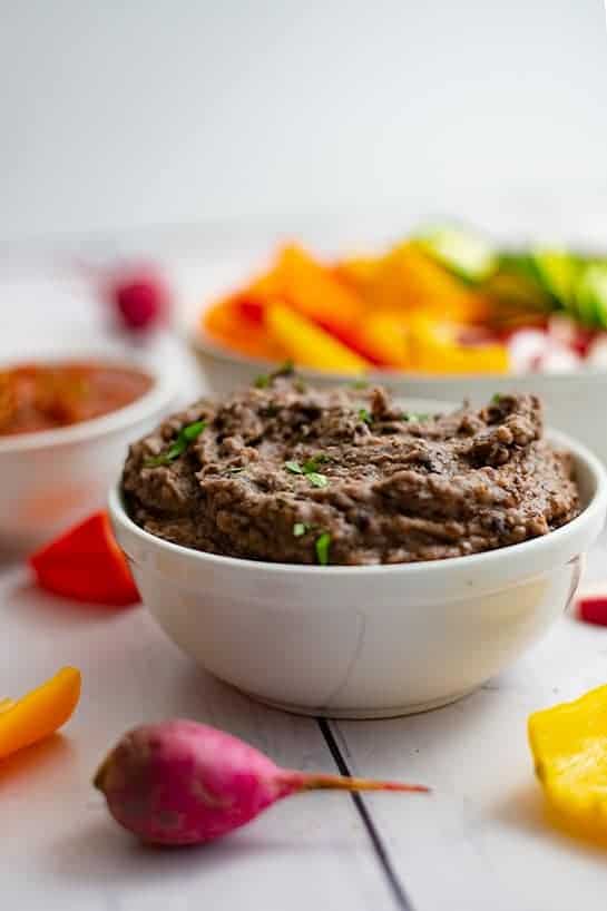 Small white bowl of 3 ingredient black bean dip recipe with radish in front of the bowl and a bowl of sliced vegetables in the background