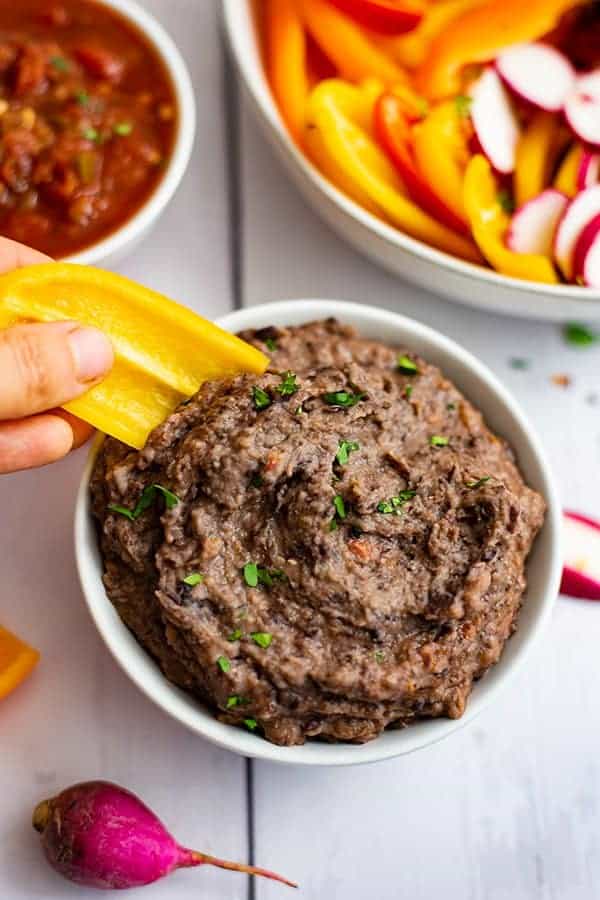 Yellow mini pepper dipped in a bowl of black bean dip recipe with salsa and chopped vegetables in the background
