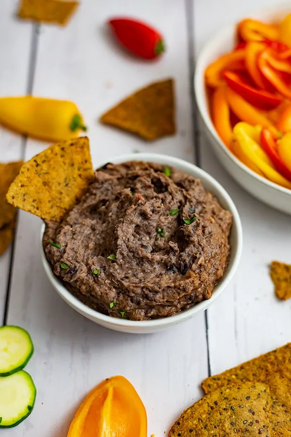Bowl of the 3 ingredient black bean dip recipe with a chip in the dip with veggies in the background