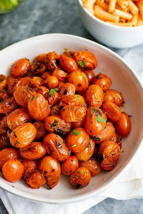 Close up shot of a large white bowl of blistered tomatoes, topped with fresh parsley and a small bowl of lentil pasta in the background