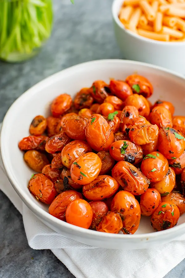 White bowl of blistered tomatoes over a white napkin with a small bowl of lentil pasta in the background