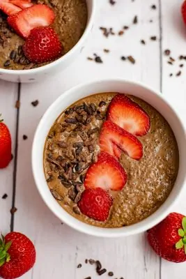 Overhead shot of a white bowl filled with breakfast quinoa bowl recipe, with second bowl in the background and surrounded by strawberries and cacao nibs.
