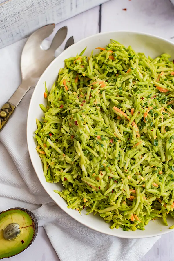 This large bowl of broccoli slaw salad with avocado ranch dressing in a white bowl on a white napkin with a large serving spoon behind