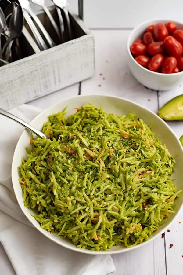 This large bowl of broccoli slaw salad with avocado ranch dressing in a white bowl on a white napkin with fork in the bowl and a bowl of tomatoes to the right