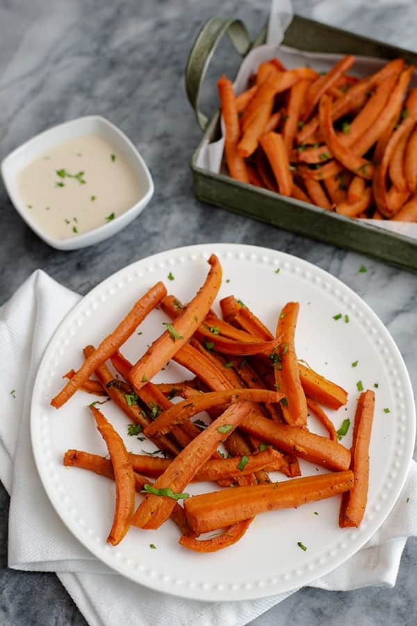 Overhead shot of spicy curry oven baked carrot fries on a white plate and a white napkin with a small ramekin of lime cilantro tahini sauce in the background