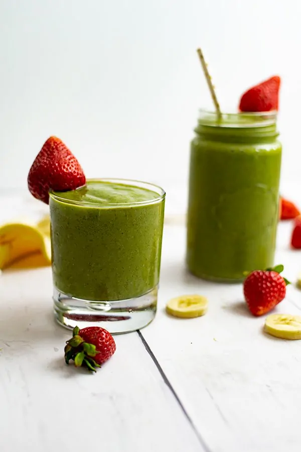 Two glasses of the strawberry banana green smoothie, one with a straw in the glass, surrounded by strawberries and banana 