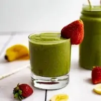 A Small glass full of strawberry banana green smoothie with a strawberry on the rim with a cut up banana in the background as well as a larger glass of green smoothie