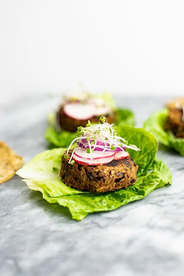 Vegan Black bean burger recipe over lettuce leaves topped with sliced radish, red onion and sprouts on a marble slab with burgers in the background.