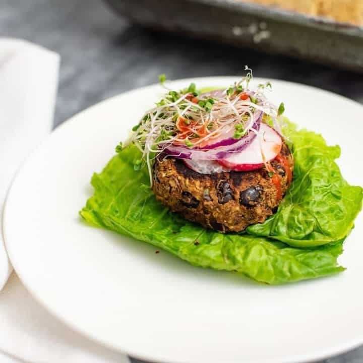 Vegan Black bean burger recipe over lettuce leaves topped with sliced radish, red onion and sprouts on a white plate.