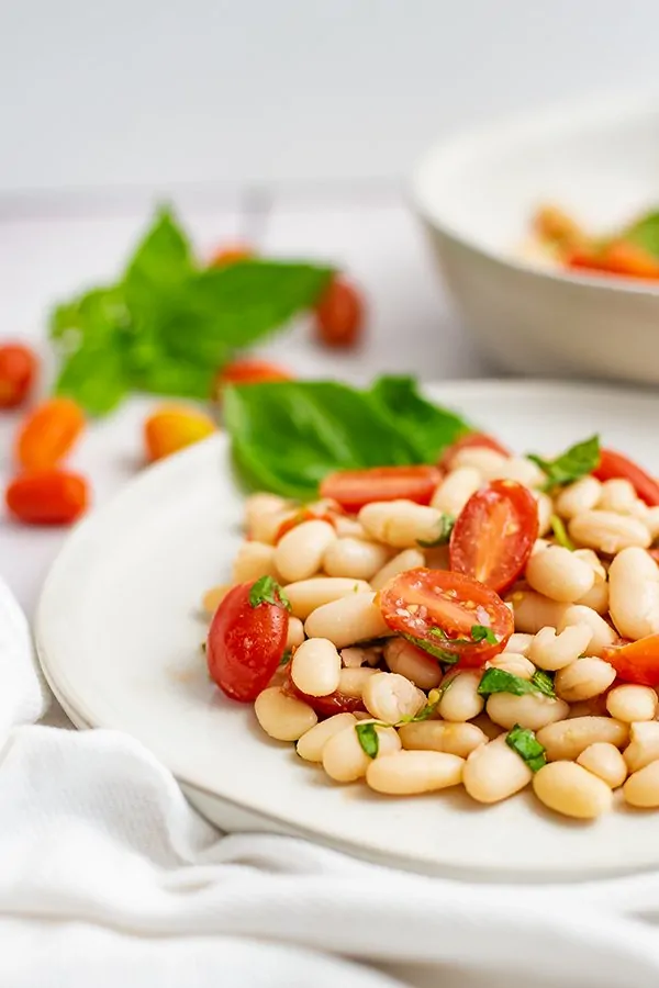 Vegan cannellini bean salad on a white plate with a white napkin with basil and tomatoes in the background 