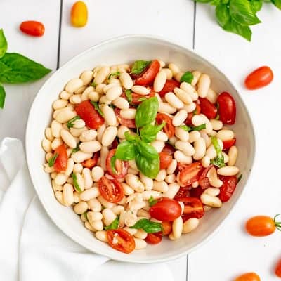 Overhead shot of bowl of vegan cannellini bean salad in a white bowl with tomatoes and basil around the plate.