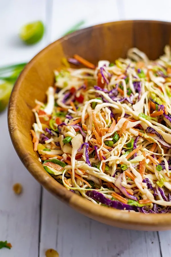 Large wooden salad bowl filled with Asian Cabbage slaw surrounded by peanuts and limes. 