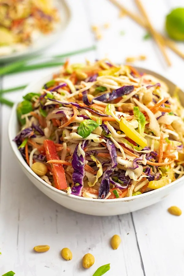 Large white bowl filled with Asian Slaw with peanut dressing surrounded by peanuts and green onions. 