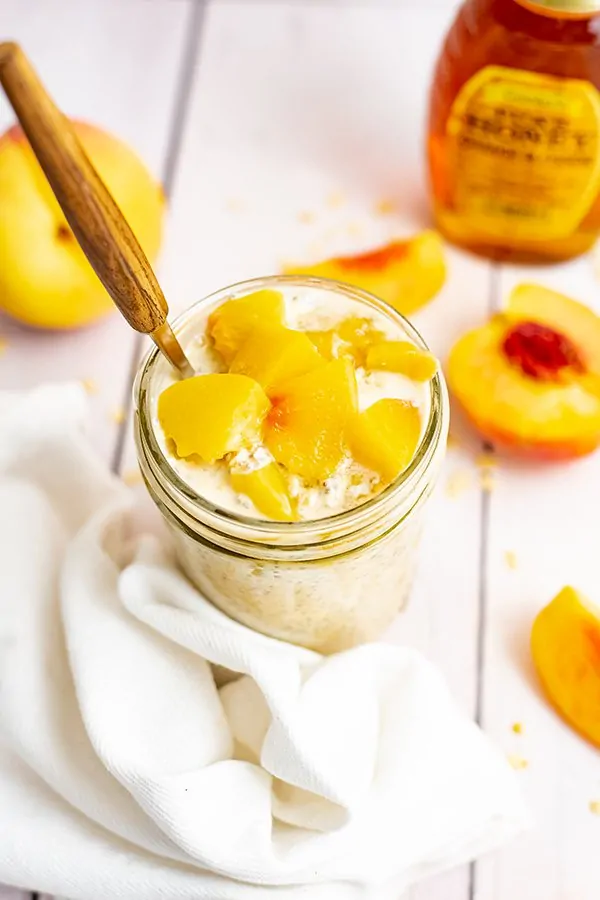 Overhead shot of Small mason jar filled with peach overnight oats with yogurt with sliced peaches on top with a wooden handle spoon in the jar.