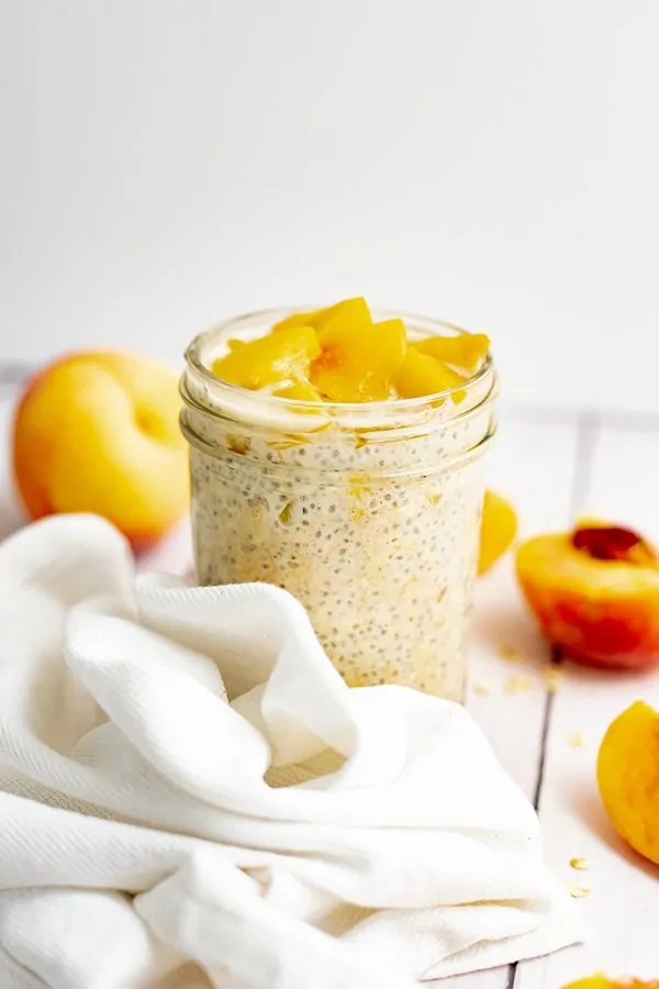 Small mason jar filled with peach overnight oats with yogurt with sliced peaches on top surrounded by a white napkin and sliced peaches