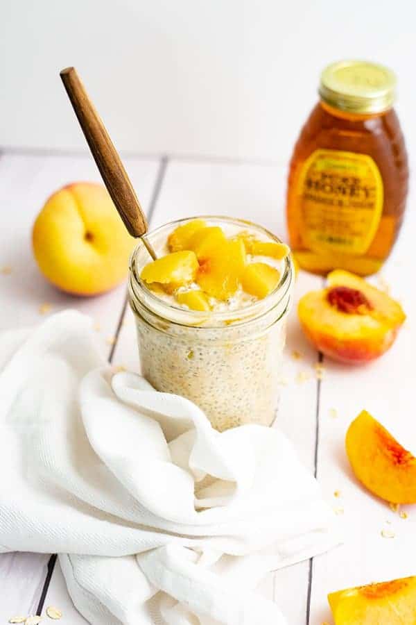 Small mason jar filled with peach overnight oats with yogurt with sliced peaches on top with a brown handled spoon in the jar and a peach in the background