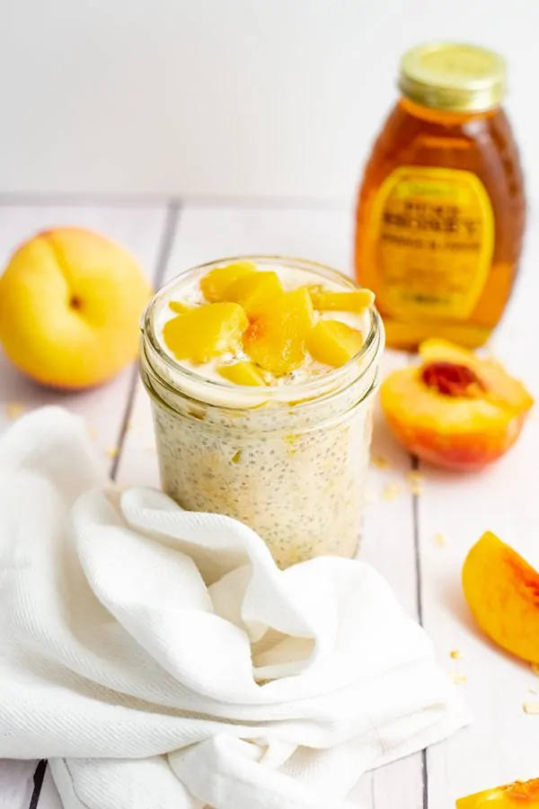 Small mason jar filled with peach overnight oats with yogurt with sliced peaches on top surrounded by sliced peaches and a bottle of honey.