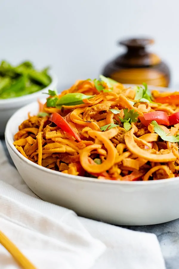 Close up shot of large white bowl filled with spicy peanut spiralized sweet potato pasta with a small brown vase in the background. 