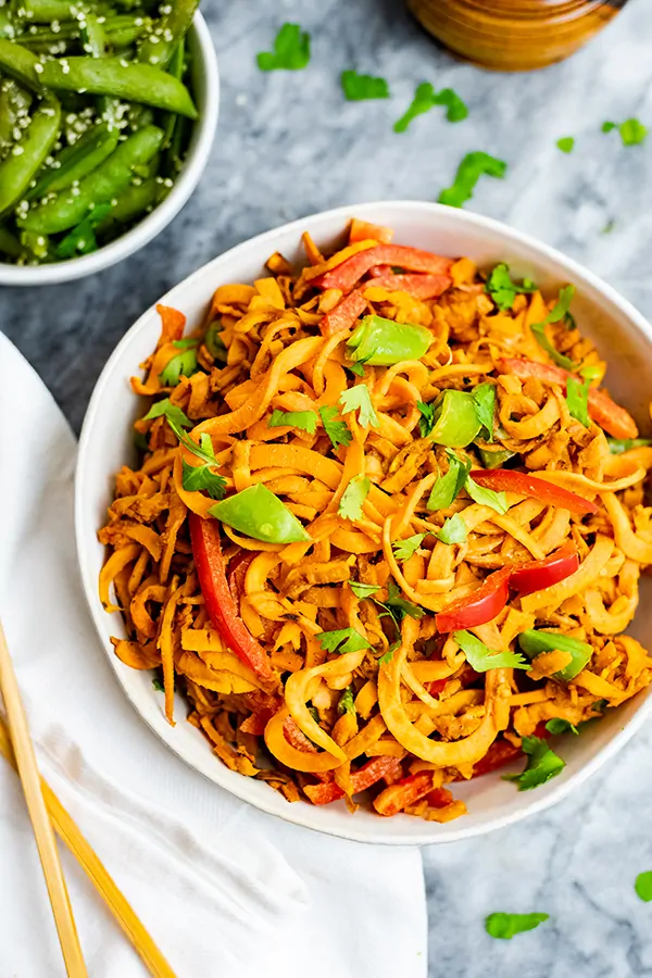spicy peanut spiralized sweet potatoes in a large white bowl with chopsticks to the left side and green snap peas in the background to the left