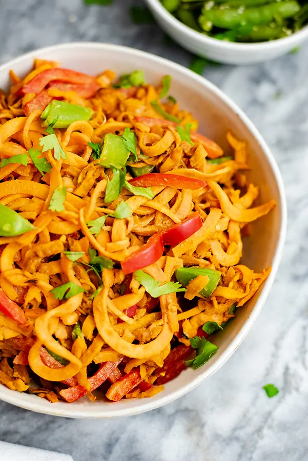 Overhead shot of a large white bowl filled with spicy peanut spiralized sweet potato pasta topped with cilantro with a bowl of snap peas in the background to the right.