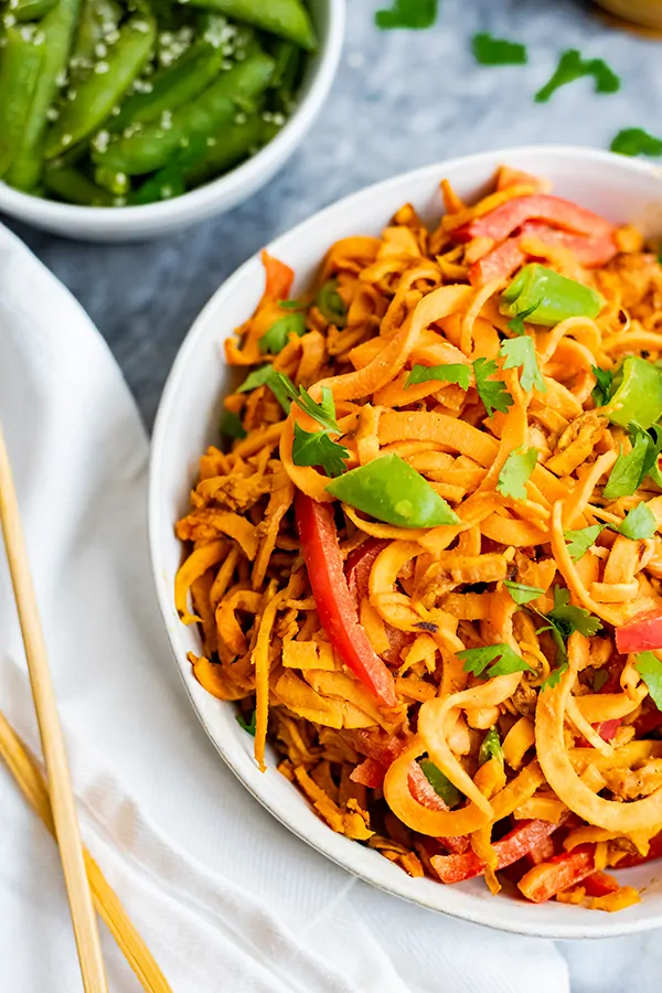 Overhead shot of spicy peanut spiralized sweet potatoes in a large white bowl with chopsticks to the left side and green snap peas in the background