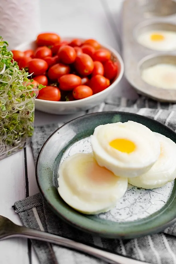 Three perfectly poached eggs in the oven on a blue plate over a blue striped napkin with a fork in front of the plate and a bowl of tomatoes and a muffin tin filled with poached eggs in the background