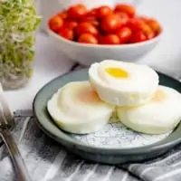 Three perfectly poached eggs in the oven stacked on top of each other on a blue plate and a blue striped nakpkin with sprouts and tomatoes in the background